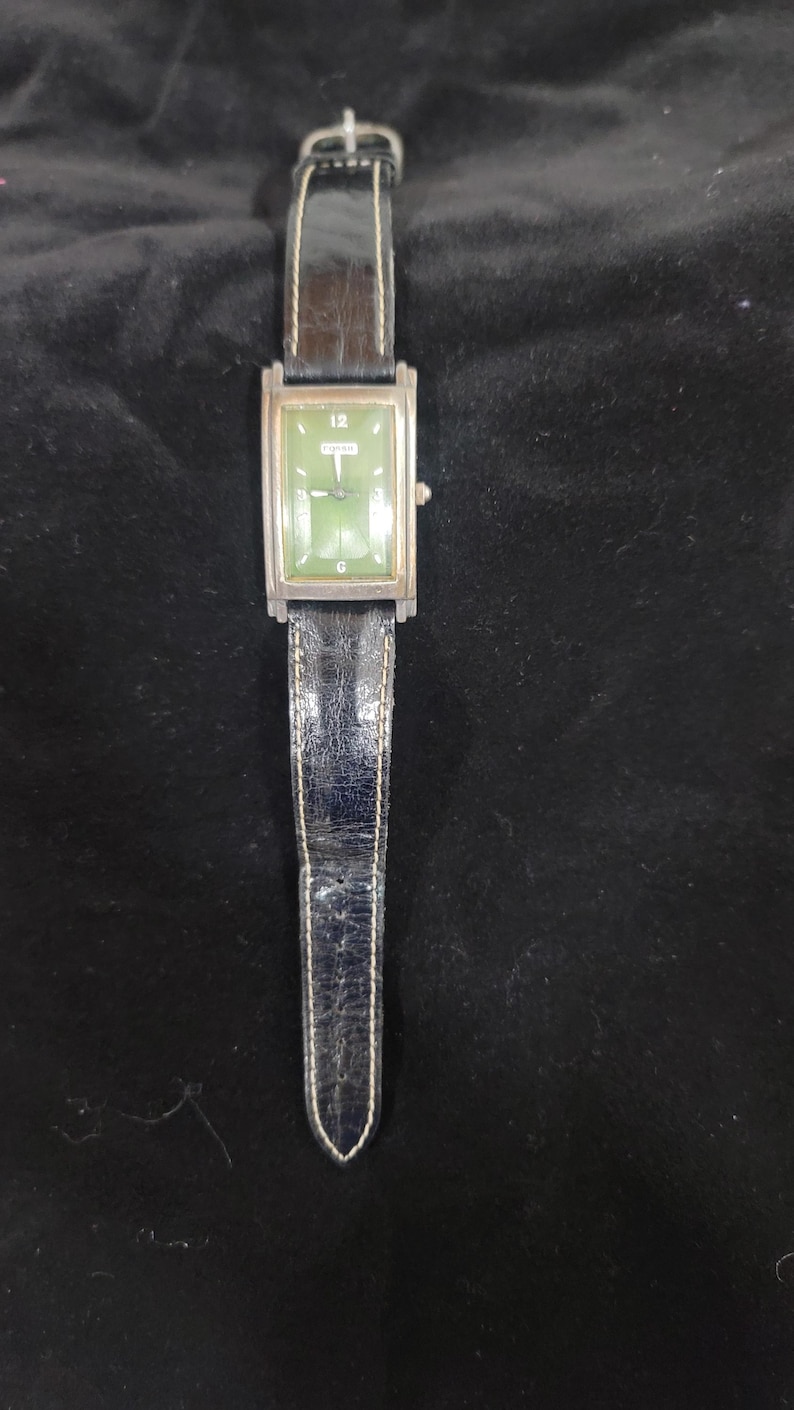 May include: A rectangular wristwatch with a green face, silver-tone metal casing, and a black leather strap with white stitching. The watch face features the number 12 at the top, the Fossil logo in the center, and a G at the bottom position.