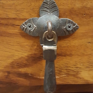 May include: A black metal drawer pull with a leaf design and a hook. The pull is attached to a wooden surface.