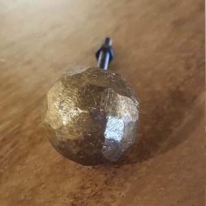 Puede incluir: Un pomo redondo texturizado de color dorado con un vástago de metal negro. El pomo está hecho de un metal rugoso y martillado.