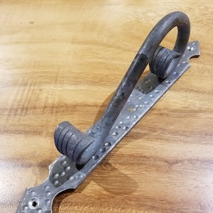 May include: A silver metal door handle with a hammered finish and a decorative scroll design. The handle is attached to a long, rectangular plate with a pointed end.