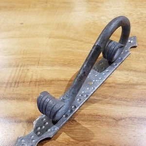 May include: A silver metal door pull with a hammered finish and a decorative scroll design. The pull is mounted on a rectangular plate with a pointed end.