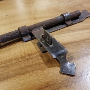 May include: An aged metal bolt latch, featuring a sliding bar and mounting plate. The latch shows signs of wear, with a dark, weathered appearance. The design is simple and functional, suggesting an antique or vintage origin. The metal has a rustic finish.
