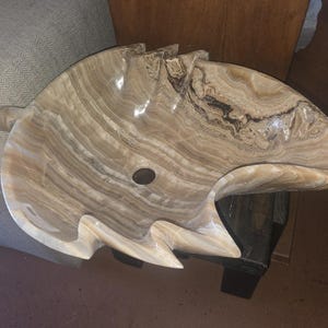 May include: A carved stone sink with a unique, organic shape. The sink is made of a light brown and cream-colored stone with dark brown veining. It features a central drain hole and a curved, wave-like design.