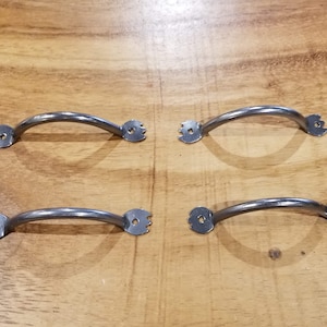 Può includere: Quattro maniglie per armadi in metallo argentato con un design curvo e un bordo decorativo. Le maniglie sono disposte in due file di due.