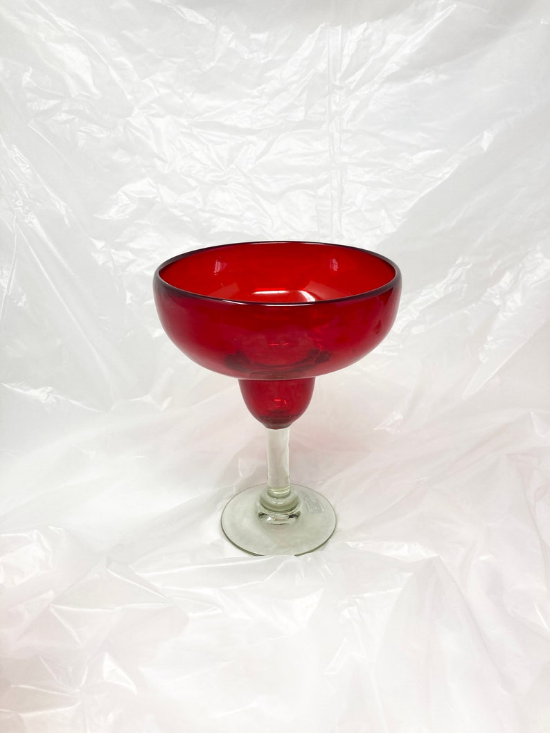 May include: A red glass margarita glass with a clear glass stem and base. The glass is empty and sitting on a white surface.