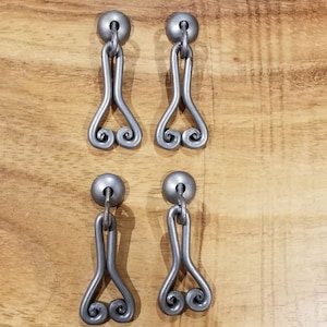 May include: Four silver metal drawer pulls with a decorative scroll design. Each pull has a round knob at the top and a long, narrow body that curves out at the bottom.