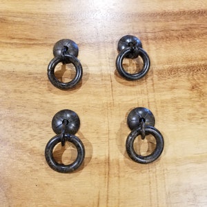 Könnte beinhalten: Vier schwarze Metall-Ringgriffe mit dekorativem Design. Die Griffe haben eine runde Basis und einen Ring, der mit einer kleinen Kette an der Basis befestigt ist.