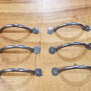 Könnte beinhalten: Sechs silberne Metall-Schubladengriffe mit gebogenem Design und dekorativem Rand. Die Griffe sind in zwei Reihen zu je drei angeordnet.