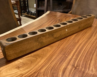May include: A long, rectangular wooden candle holder with ten evenly spaced circular openings. The holder is made of light brown wood and sits on a large, wooden table. The background includes other wooden furniture and decorative items.