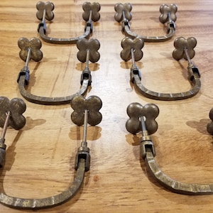 Pode incluir: Dez puxadores de armário de latão antigo com um design de trevo de quatro folhas. Cada puxador tem uma alça em forma de U e um acabamento decorativo e ornamentado.