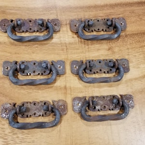 Peut inclure: Six poignées de tiroir de style antique avec un design en métal torsadé. Les poignées sont en métal foncé avec une finition rustique et ont une base rectangulaire avec des détails ornés.