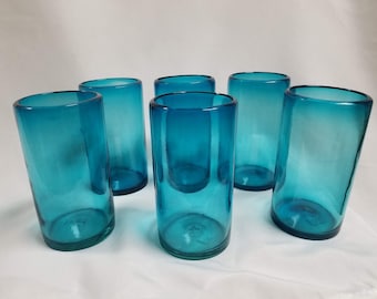 May include: Six turquoise blue glass tumblers, each with a slightly different shade of blue, are arranged in a group on a white surface.