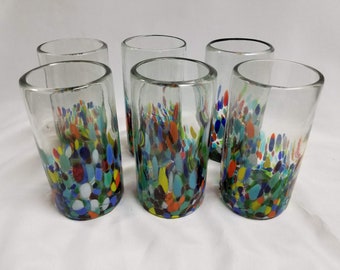 May include: Six clear glass tumblers with a colorful, speckled design. The pattern includes blue, green, yellow, orange, and red spots. The glasses are cylindrical and appear to be handmade, perfect for serving beverages.