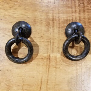 May include: Two antique-style cabinet pulls with a dark bronze finish. Each pull features a round backplate and a circular ring handle. The hardware is mounted on a light-colored wooden surface, creating a contrast in textures and tones.