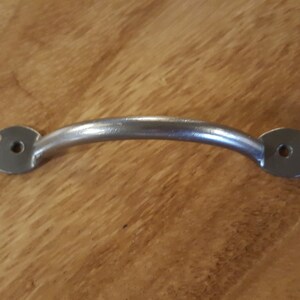 Pode incluir: Uma puxador de armário de metal prateado com uma alça curva e extremidades decorativas. A alça é lisa e as extremidades têm um design festoneado.