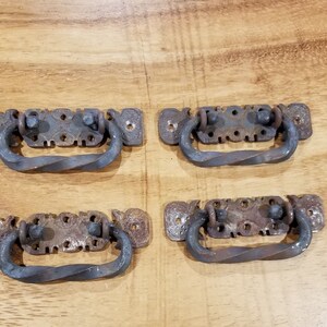 Peut inclure: Quatre poignées de tiroir en fer de style antique avec un design torsadé. Chaque poignée a une base rectangulaire avec des détails décoratifs et une poignée en métal torsadée.