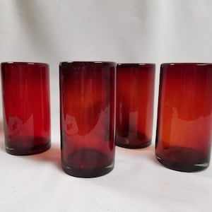 May include: Four red glass tumblers, each with a slightly different shade of red, are arranged on a white surface. The glasses are all the same size and shape, and they have a smooth, rounded bottom.