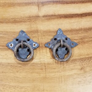 May include: Two antique-style metal door pulls with a star-shaped design and a ring handle. The pulls are a dark gray color and have a rustic finish.