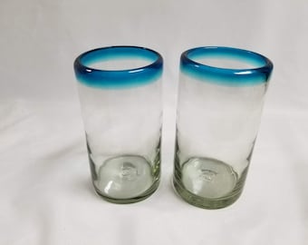 May include: Two clear glass tumblers with a blue rim. The glasses are empty and sitting on a white surface.