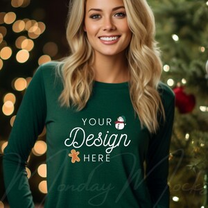 May include: A woman wearing a dark green long-sleeved shirt with the text "YOUR Design HERE" and a gingerbread man and snowman graphic. The shirt is in front of a Christmas tree with lights.