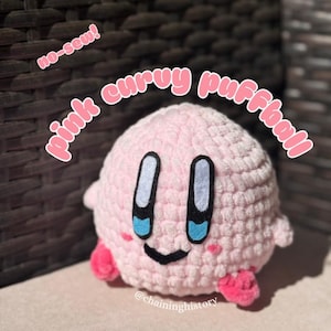 以下が含まれることがあります： ピンク色の、かぎ針編みのパフボールのおもちゃ。大きな黒と白の目、青いアクセント、小さなピンク色の足が付いています。「pink curvy puffball」の文字はピンク色の筆記体で書かれています。「no-sew!」の文字はピンク色の筆記体で書かれています。手作りです。