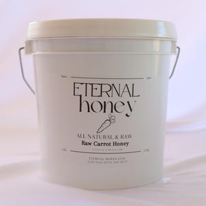 May include: A white plastic bucket with a black label that reads "ETERNAL honey" and "ALL NATURAL & RAW Raw Carrot Honey". The label also includes a small carrot illustration and the company's website and phone number.