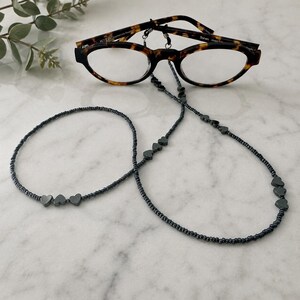 May include: Tortoiseshell-patterned spectacles attached to a long beaded chain. The chain consists of small, dark metallic-coloured seed beads interspersed with clusters of three small, dark heart-shaped beads at regular intervals along the length.