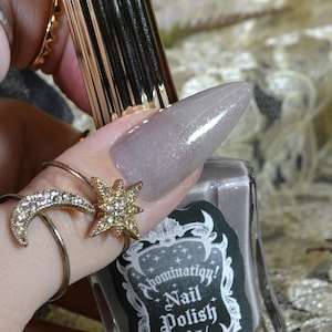 May include: A clear glass bottle of gray nail polish with a gold cap. The bottle label reads "Domination! Nail Polish 11.8 ml / 1/2 fl oz".