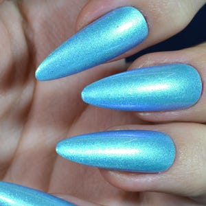 Op de afbeelding: Close-up van een hand met lange, puntige, iriserende blauwe nagels. De nagels hebben een glinsterende, parelmoerachtige afwerking die licht reflecteert. Een gouden ring is zichtbaar op de ringvinger.