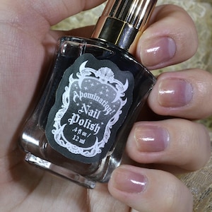 May include: A clear glass bottle of black nail polish with a gold cap. The bottle has a white label with the text "Apothecary Nail Polish 4ml/ 1/2 fl oz".