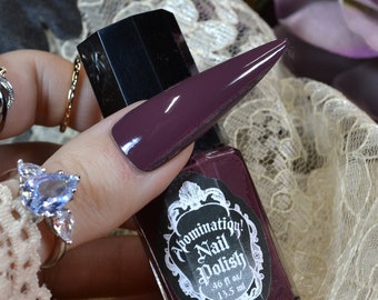 Könnte beinhalten: Eine Flasche Abomination! Nagellack in einem tief burgunderroten Farbton, die gehalten wird, um die Farbübereinstimmung mit einem langen, spitzen Nagel zu zeigen. Die Flasche hat einen schwarzen Deckel und ein Etikett mit verziertem Text. Der Hintergrund umfasst Spitze und florale Elemente.