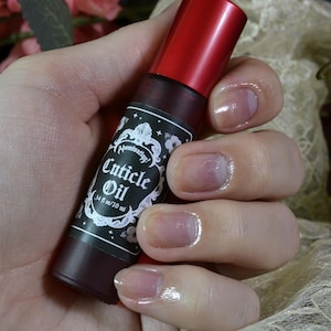 Op de afbeelding: Een rode en zwarte fles nagelriemolie met een wit label waarop "Cuticle Oil" en "3.4 fl oz/10 ml" staat.
