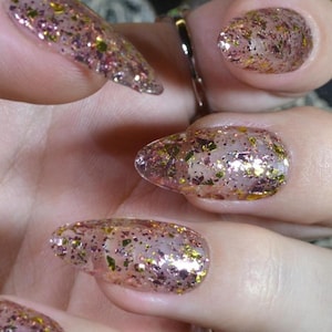 May include: Close-up of a hand with long, glittery, pink and gold nail polish. A black ring with a large, clear stone sits on the ring finger. The nails are a light pink base with gold and pink glitter. The background includes lace and flowers.