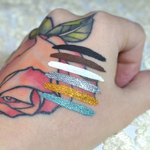 May include: A hand displays six swatches of metallic and matte eyeshadow colors. The shades include black, brown, white, silver, gold, and teal. The hand has a floral tattoo.