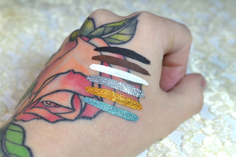 May include: A hand displays six swatches of metallic and matte eyeshadow colors. The shades include black, brown, white, silver, gold, and teal. The hand has a floral tattoo.