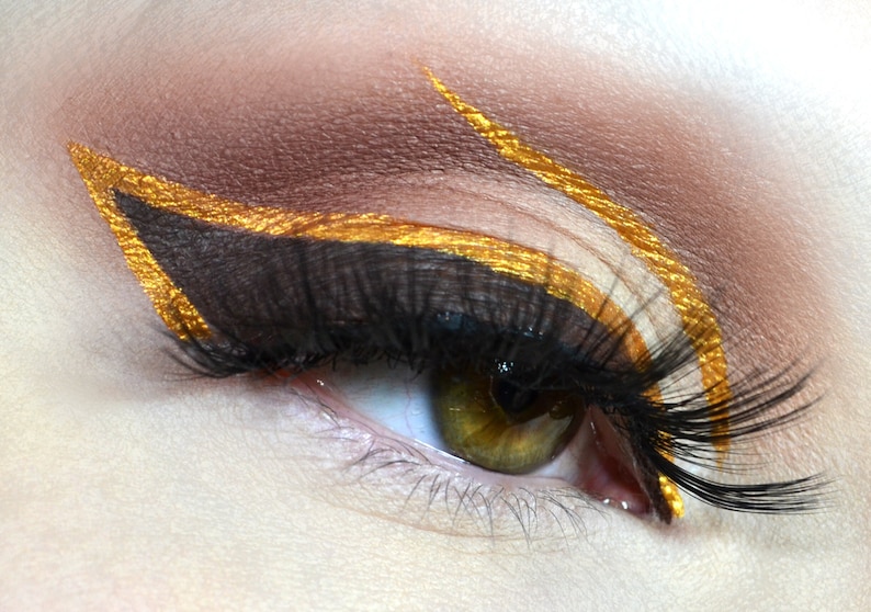 May include: Close-up of an eye with dramatic makeup. The eye features a brown eyeshadow base with gold eyeliner outlining the upper lid and extending outwards. Long, black eyelashes and a green iris complete the look.