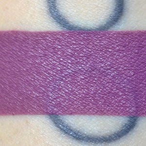 Puede incluir: Una muestra horizontal gruesa de pigmento morado profundo mate aplicado directamente sobre la piel. La textura del producto cosmético parece suave y opaca, cubriendo la superficie de la piel de manera uniforme con un tono violeta saturado.