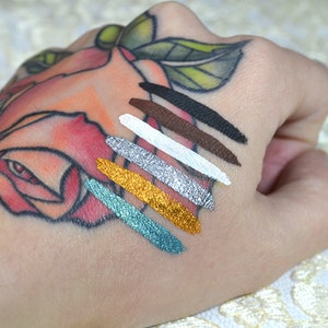 May include: A hand with a rose tattoo displays seven swatches of colorful eyeliner. The colors include black, brown, white, silver, gold, and teal. The swatches are applied in diagonal lines across the skin.
