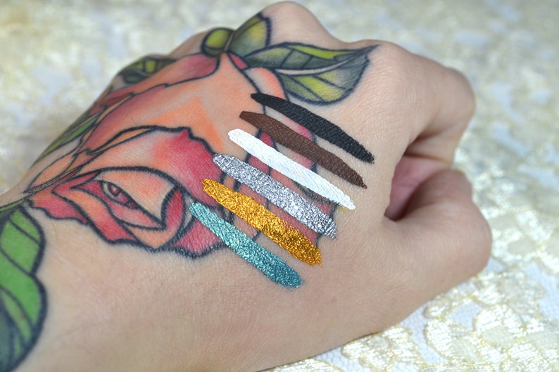 May include: A hand with a rose tattoo displays seven swatches of colorful eyeliner. The colors include black, brown, white, silver, gold, and teal. The swatches are applied in diagonal lines across the skin.