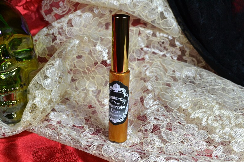 May include: A gold-colored eyeliner with a black and white label that reads "Illumination! Watercolor Liner". The eyeliner has a gold cap and is displayed on a cream-colored lace fabric.