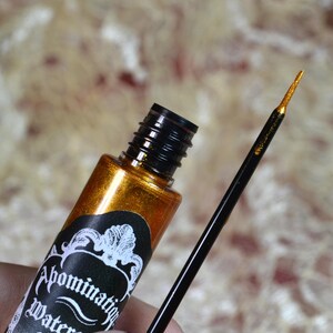 May include: A gold metallic eyeliner bottle with a black cap and applicator brush. The label reads "Abomination Water". The brush tip is gold. The background is a blurred, textured pattern.