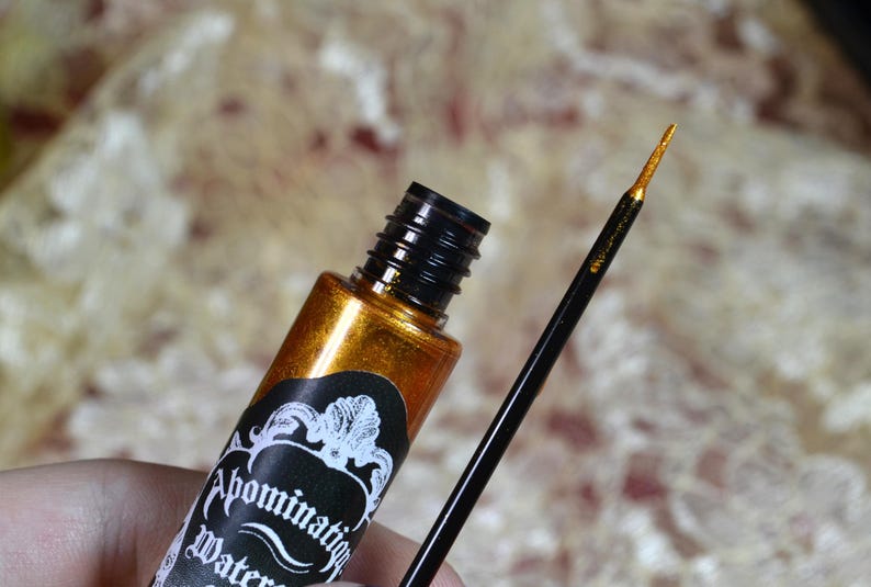 May include: A gold metallic eyeliner bottle with a black cap and applicator brush. The label reads "Abomination Water". The brush tip is gold. The background is a blurred, textured pattern.