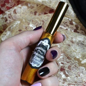 May include: A gold-colored liquid eyeliner with a gold cap is held in a hand with painted nails. The label reads "Abominatrix Watercolor Liner" with a decorative border. The bottle contains .27 fl oz / 8 ml of product.