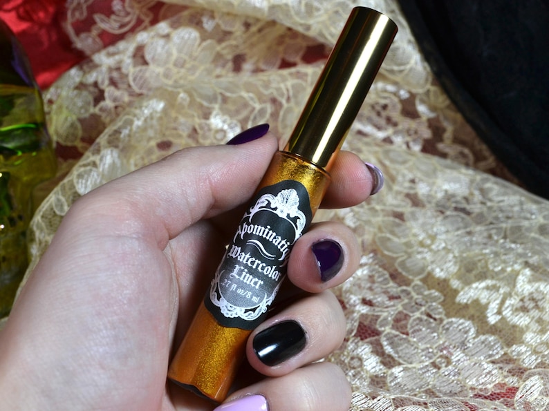May include: A gold-colored liquid eyeliner with a gold cap is held in a hand with painted nails. The label reads "Abominatrix Watercolor Liner" with a decorative border. The bottle contains .27 fl oz / 8 ml of product.