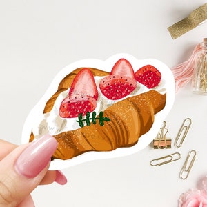 May include: A sticker featuring a croissant with whipped cream and strawberries. The sticker is on a white background and is being held by a hand.