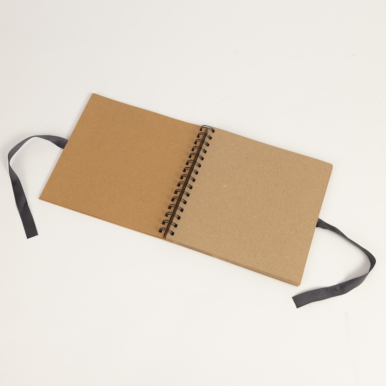 May include: A brown spiral bound sketchbook with black ribbon ties. The sketchbook is open to a blank page.