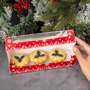 May include: A red and white cardboard box with a clear plastic window, containing three small mince pies decorated with holly leaves. The box has a snowflake design.