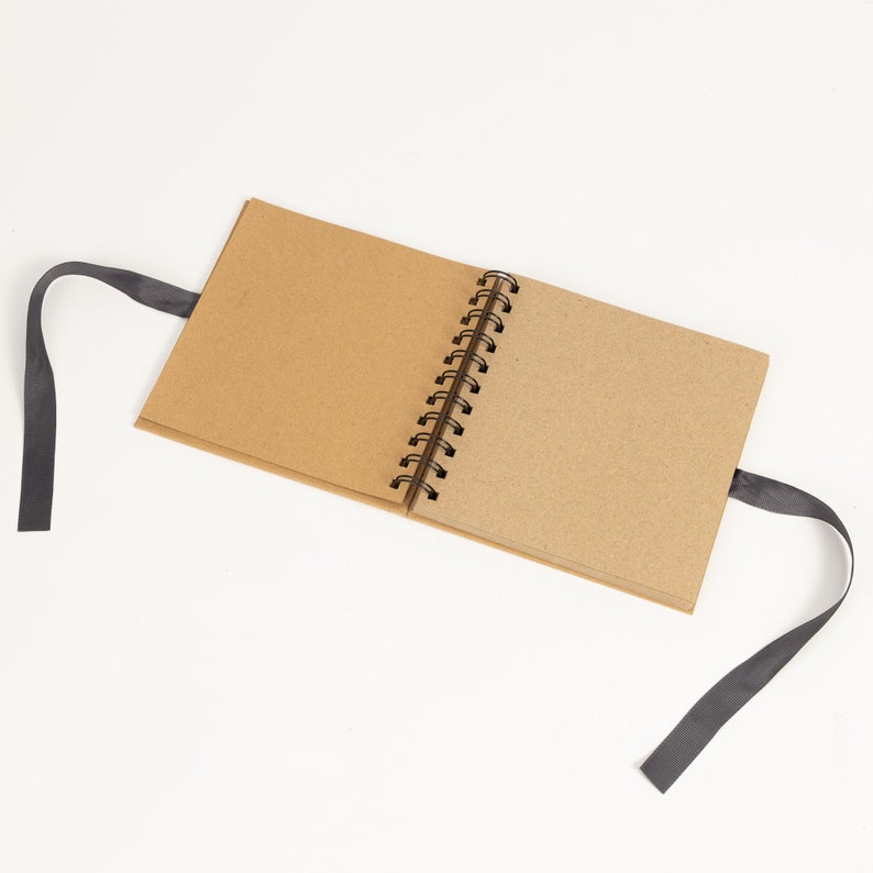May include: A brown kraft paper spiral bound notebook with a black ribbon closure. The notebook is open to a blank page.