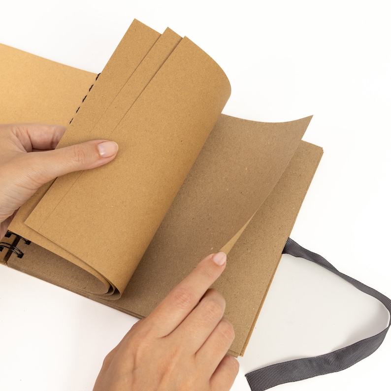 May include: A brown kraft paper journal with a black spiral binding and a gray ribbon closure. The journal is open to a blank page.
