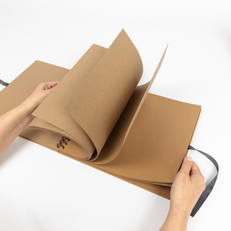 May include: A brown craft paper bound journal with a black spiral binding and a black ribbon closure. The journal is open and the pages are blank.